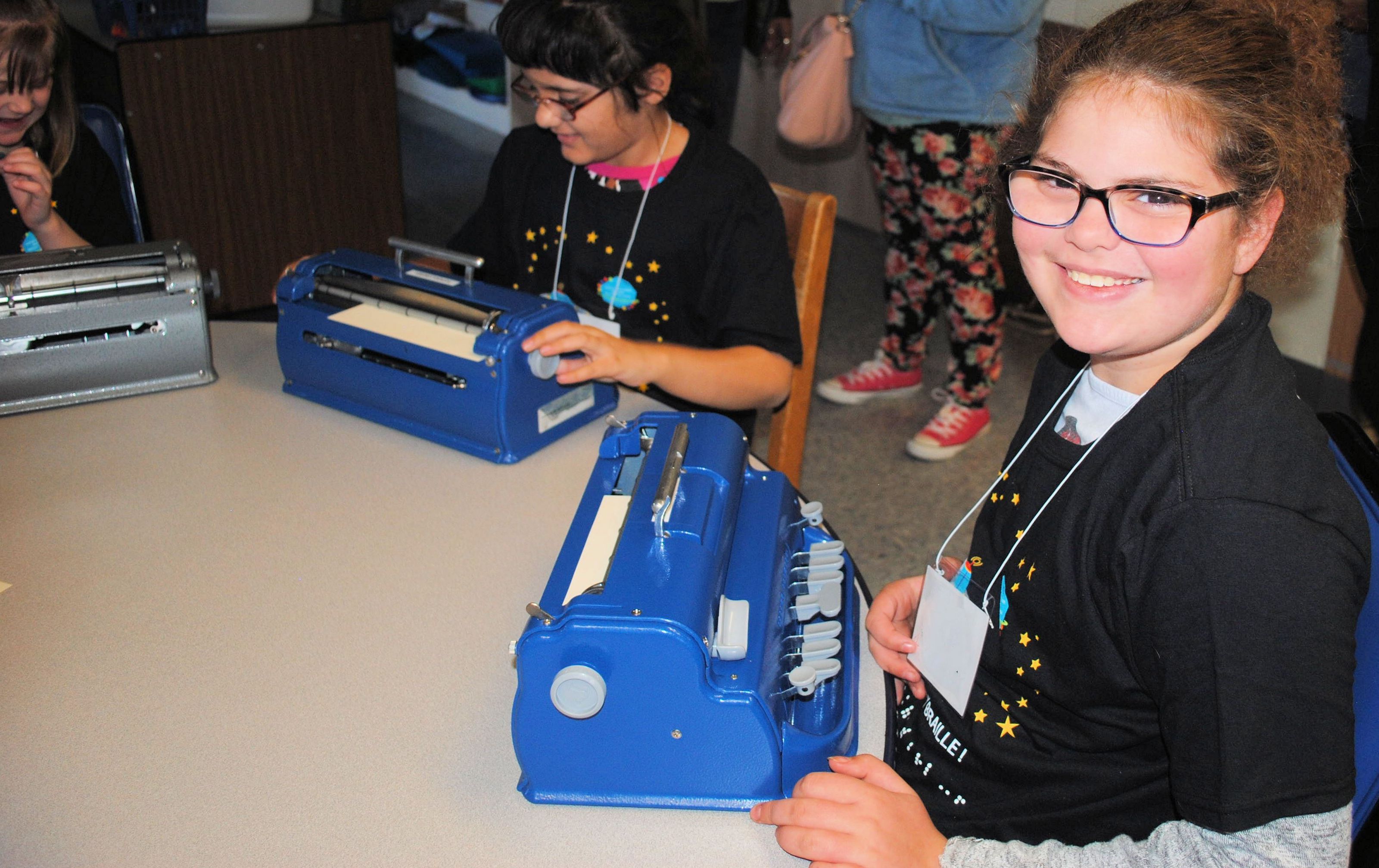 Student sitting at a table, looking at camera with a large smile. Her braille writer is on the table.