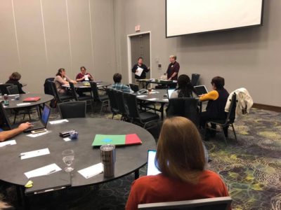 Room with people sitting around several round tables with papers and folders. A presenter standing at the front of the room.