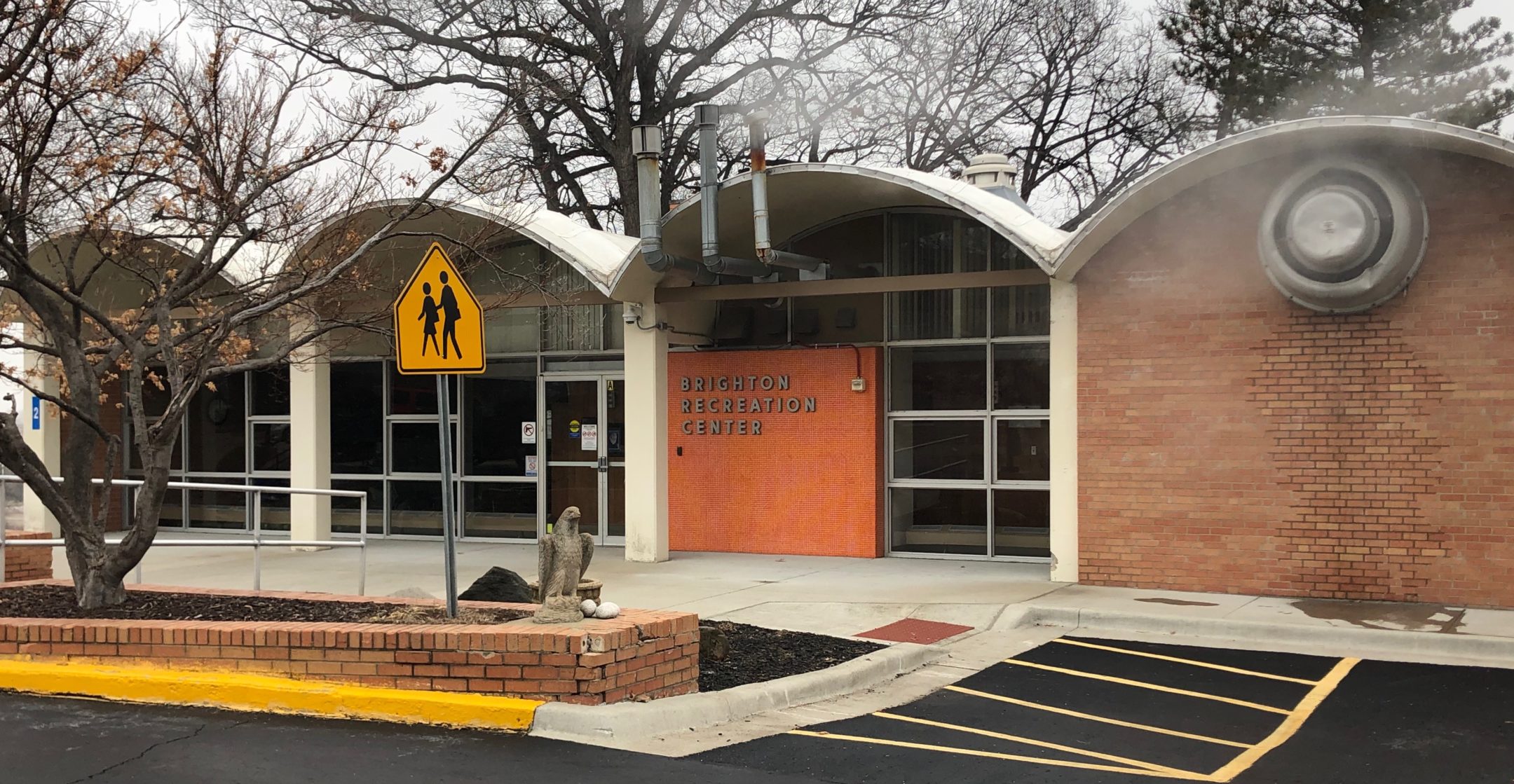 A building with 4 archway style awnings attached to white pillars. Most of the font wall is made with top to bottom windows. Under one awning is red brick wall with a heater vent creating fog into the air. An orange sign to the right of the entrance doors saying Brighten Recreation Center.