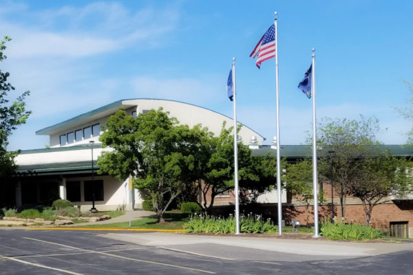 Building with a partial arch shaped space on center top with extensions to left and right. Trees on left, center and right size and 3 flags flying out in front. A parking lot in foreground of photo.