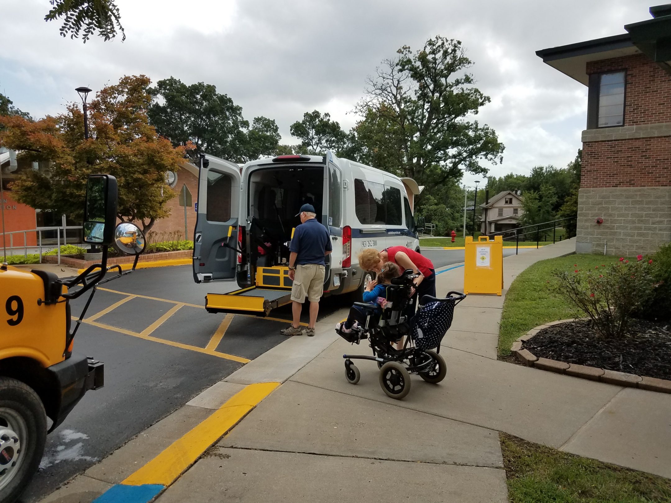 A white van with wheelchair lift parked in front of yellow cross walk between Recreation Building and Edlund Hall. A student who is in a wheelchair is waiting with teacher in red shirt to board the van. The front hood of a yellow bus is partially seen at the crosswalk.