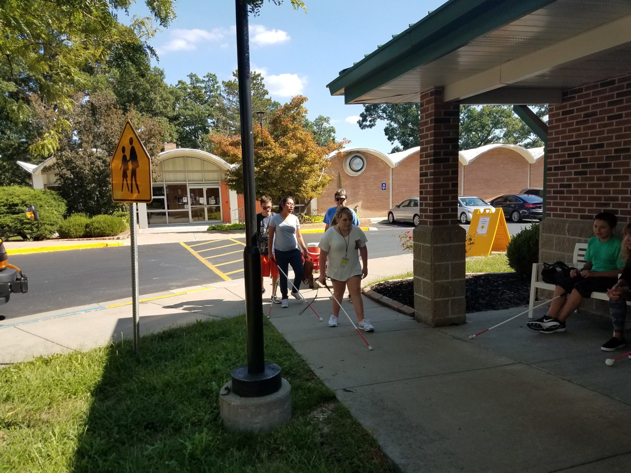 Three students arriving on campus. The recreation center in background, yellow crosswalk and then sidewalk to the back door of Edlund Hall. 2 students sitting on chairs outside the cottage as students walk by.