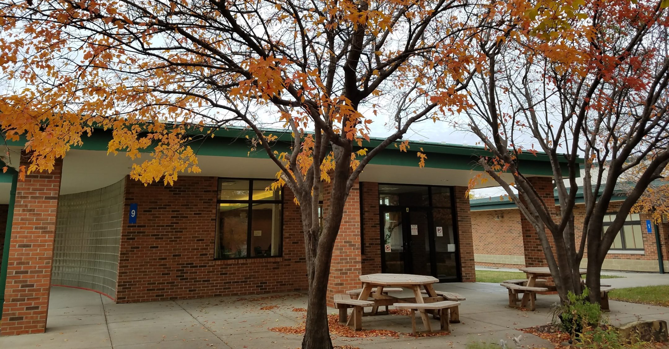 Entrance to cafeteria building showing glass opaque brick wall on the left, red brick wall with large window and a large automatic door on front facing wall. Three brick pillars and large concrete patio with two round picnic tables out front. Three large trees with leaves in fall colors out front.