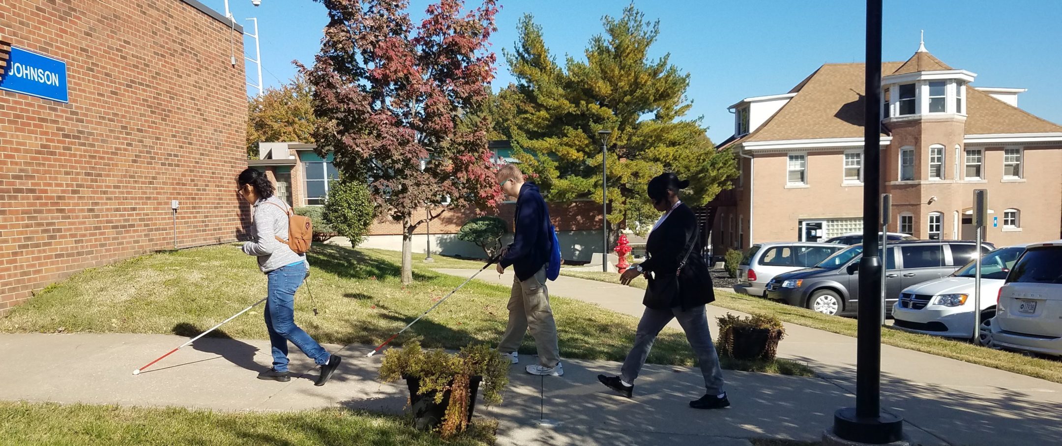 Three students walking on sidewalk with canes out front as they enter the Johnson Building. First female student wearing jeans and white shirt. The next two are males wearing coat jacket and kaki pants.