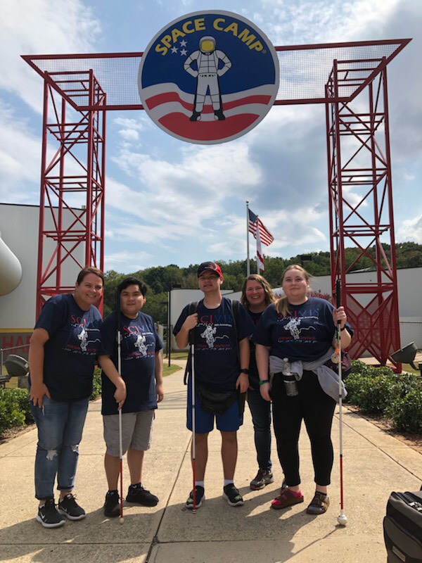 Two teachers and three students wearing spacecamp shirts and holding their canes are standing under the entrance to Space Camp.