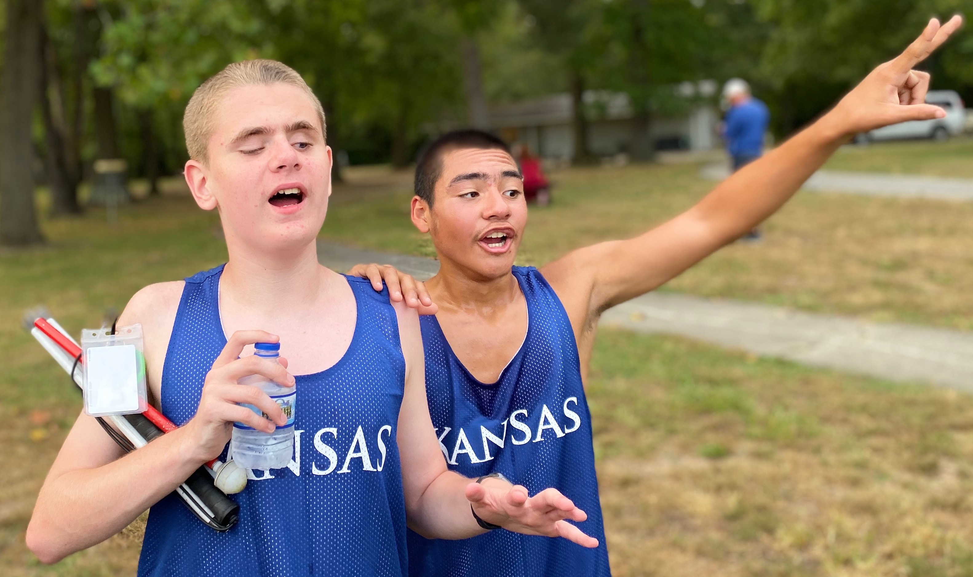 Two male students in track uniforms. One holding cane and water bottle, the other has hand on teammates shoulder and oner hand raised to the sky.