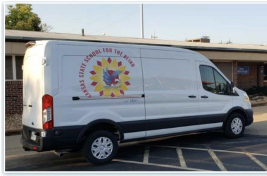 A large white van with KSSB’s new logo is parked in the circle drive, next to a display table draped with a cloth.