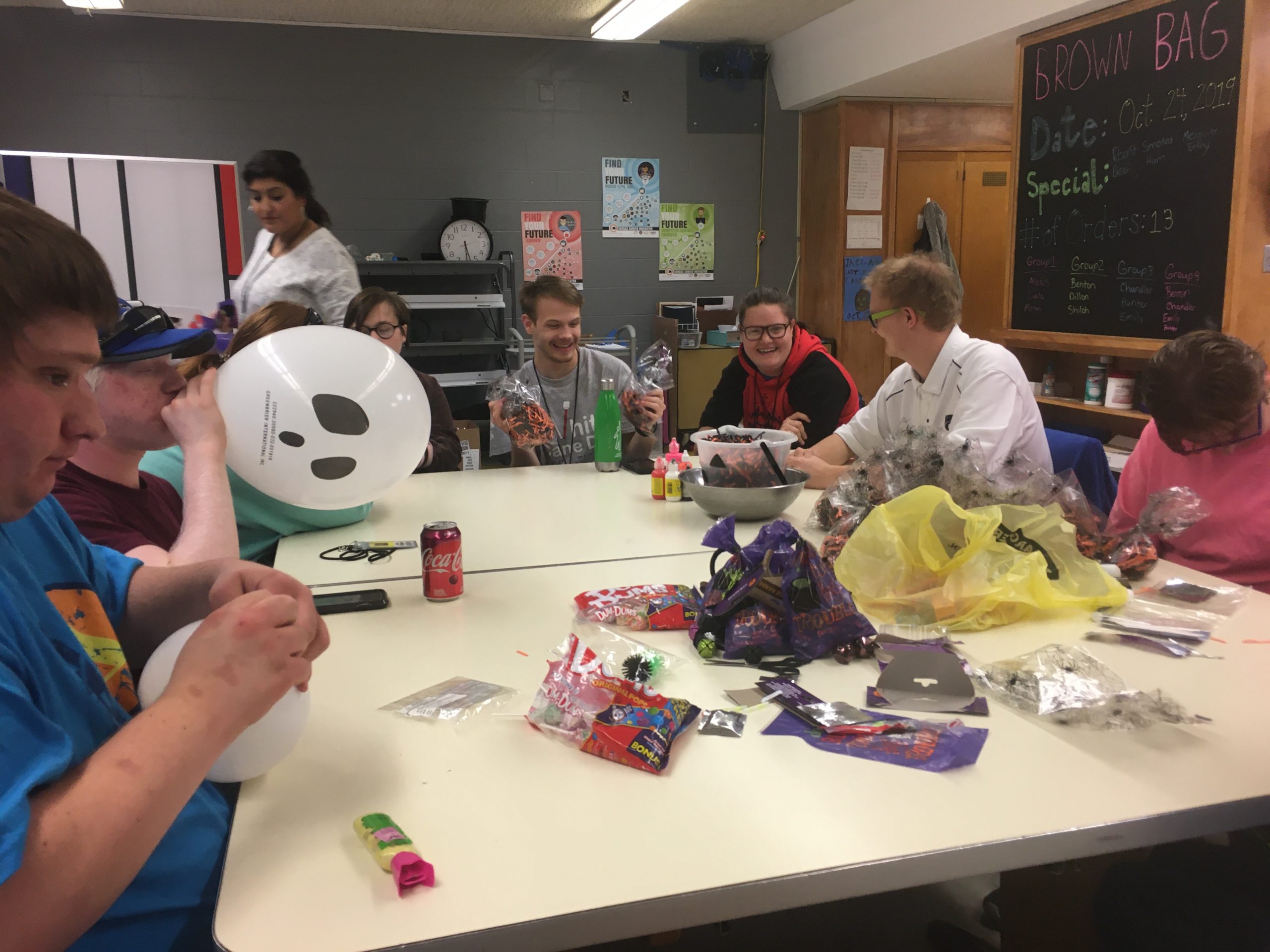 7 students sitting at a table working on a project together. One student is blowing up a balloon with a ghost face on it.