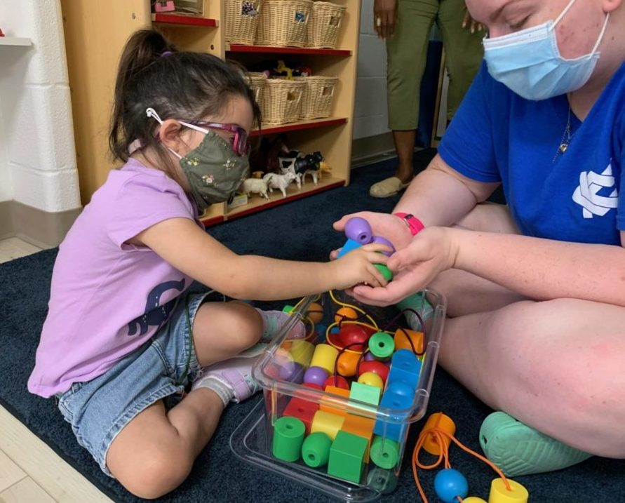 Little girl wearing glasses working with a teacher on stringing beads.