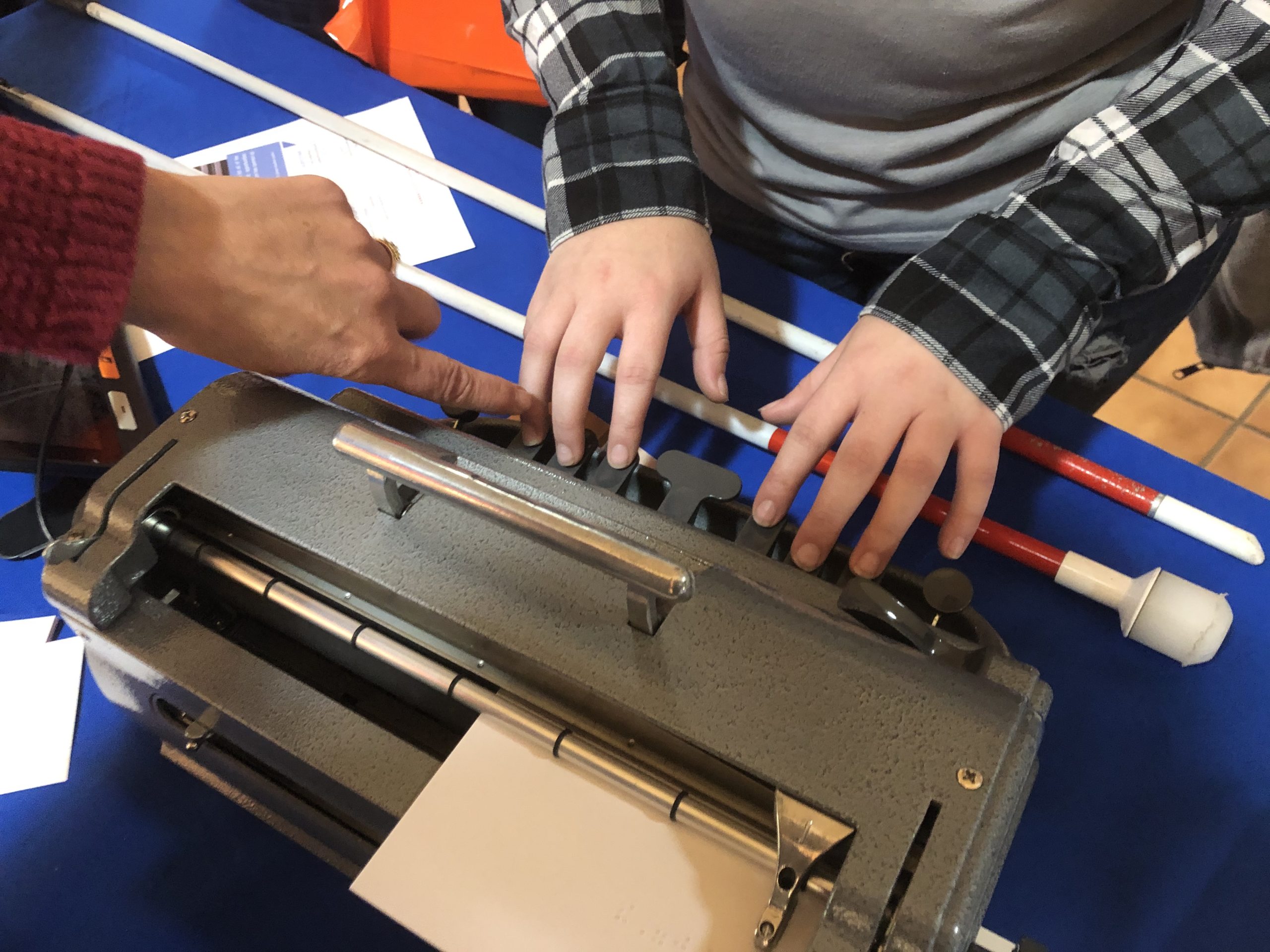 child's hands on a braillewriter and a teacher finger touching the right finger of the child's right hand. A cane rests on the table