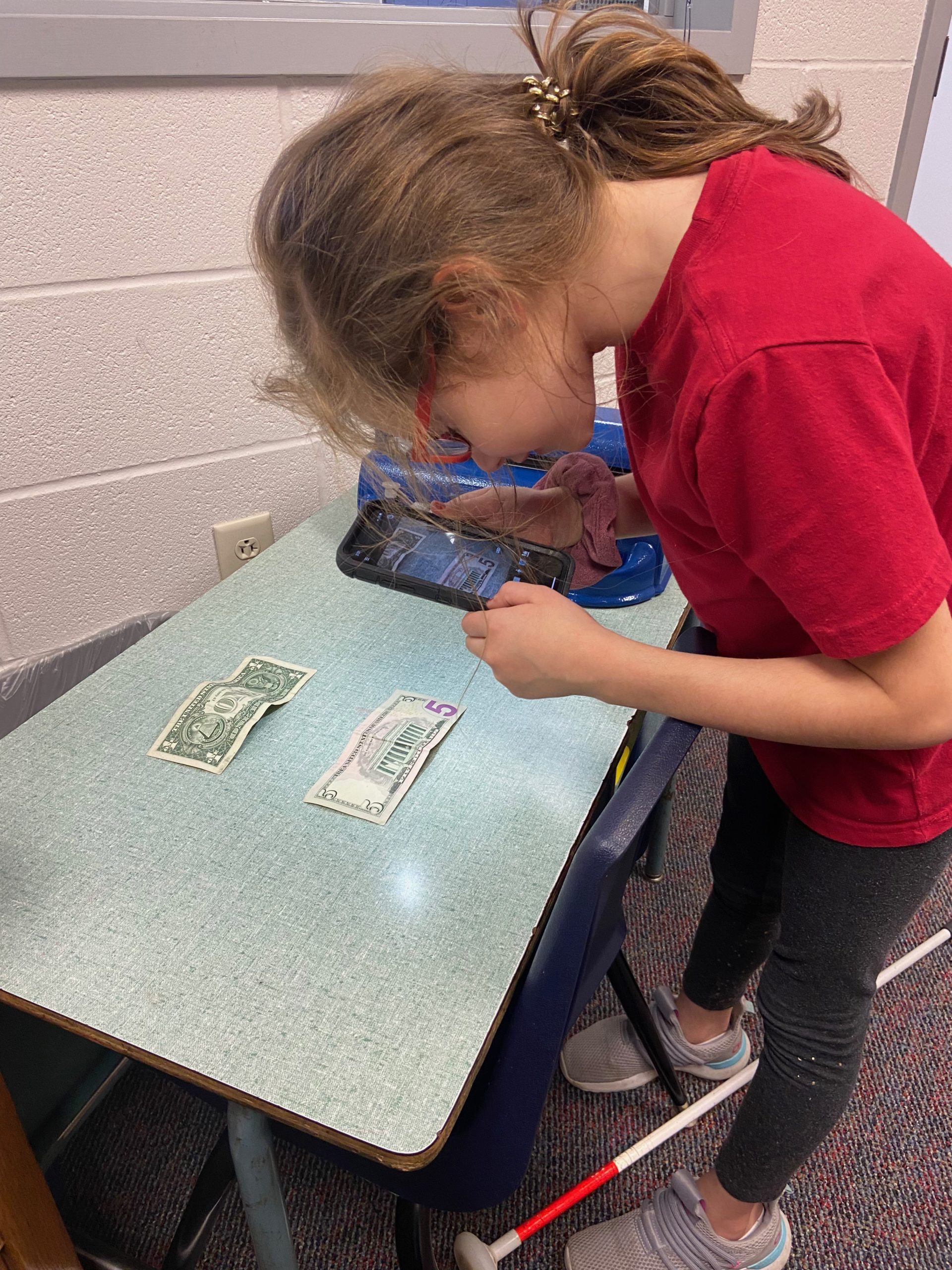 girl with glasses and brown hair holds a phone and leans over a table that a 5 dollar bill laid out