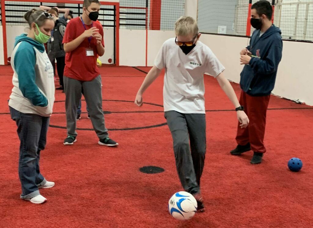 Ms. Jenny stands on the soccer field while one blond haired boy kicks a soccer ball and two other brown haired boys stand in the background talking to each other