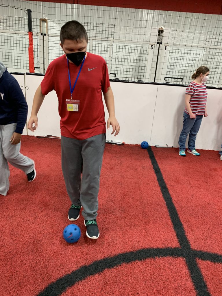 student with short hair and red shirt stand on the soccer field with a blue soccer ball in front of his right foot