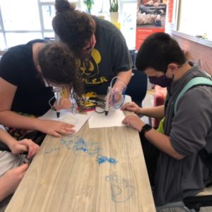 1 boy with short brown hair and 2 girls with curly brown hair pulled into a bun sit at a table together. Each has a piece of paper in front of them and they are leaning over the paper with the 3D pen