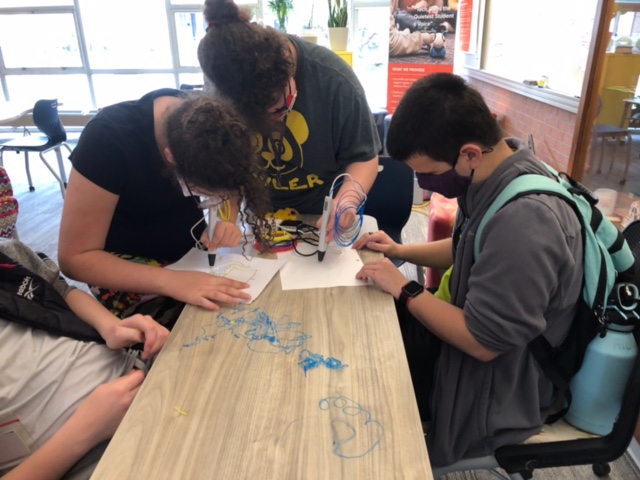 1 boy with short brown hair and 2 girls with curly brown hair pulled into a bun sit at a table together. Each has a piece of paper in front of them and they are leaning over the paper with the 3D pen