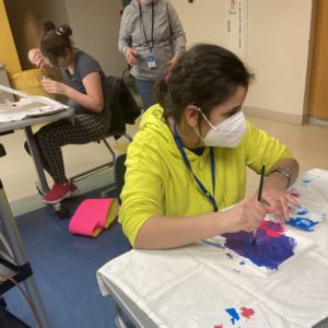 a girl sits at a table and painting on a canvas that lays flat on the table in front of her