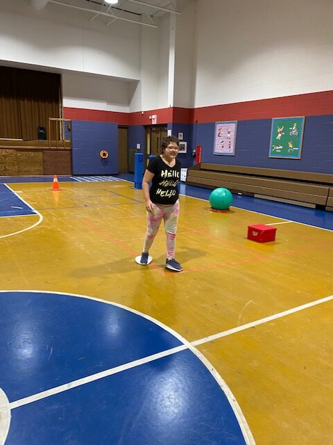 student with hair pulled back, walks down the gym floor with a paper plate under her right foot