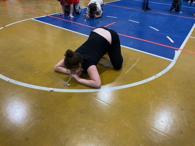 a girl with hair pulled back in the bun, uses a straw to blow a cotton ball across the gym floor