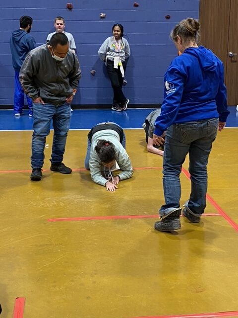 1 student on the ground using a straw to blow a cotton ball across the gym floor while 2 teachers encourage her