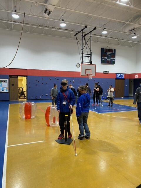 a blindfolded student, holding a cane, walks into a baseball tee that is part of an obstacle course