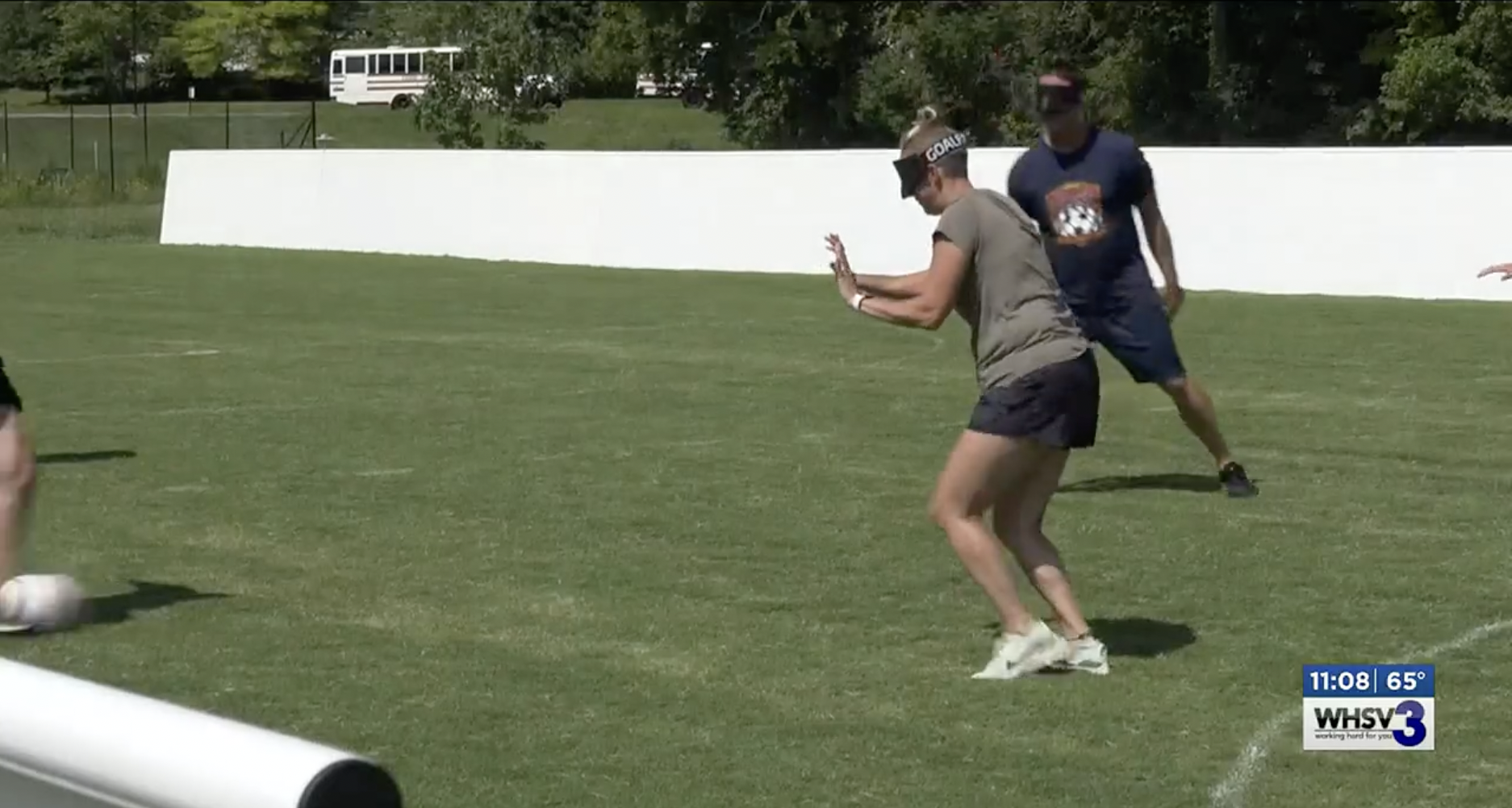 Ms. Nicole wears shorts and t-shirt and is blindfolded ready to kick a soccer ball on a soccer field