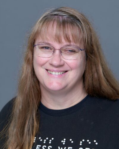 Rachel Ghram, smiling for the camera with long light brown hair and bangs and wearing glasses