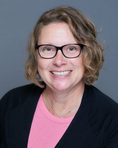 Teri Chaney smiling with short curly hair and wearing glasses.