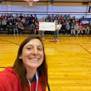 Ms. Lydia smiles while taking a selfie as a large group of students sit on gym bleachers and holds a large check