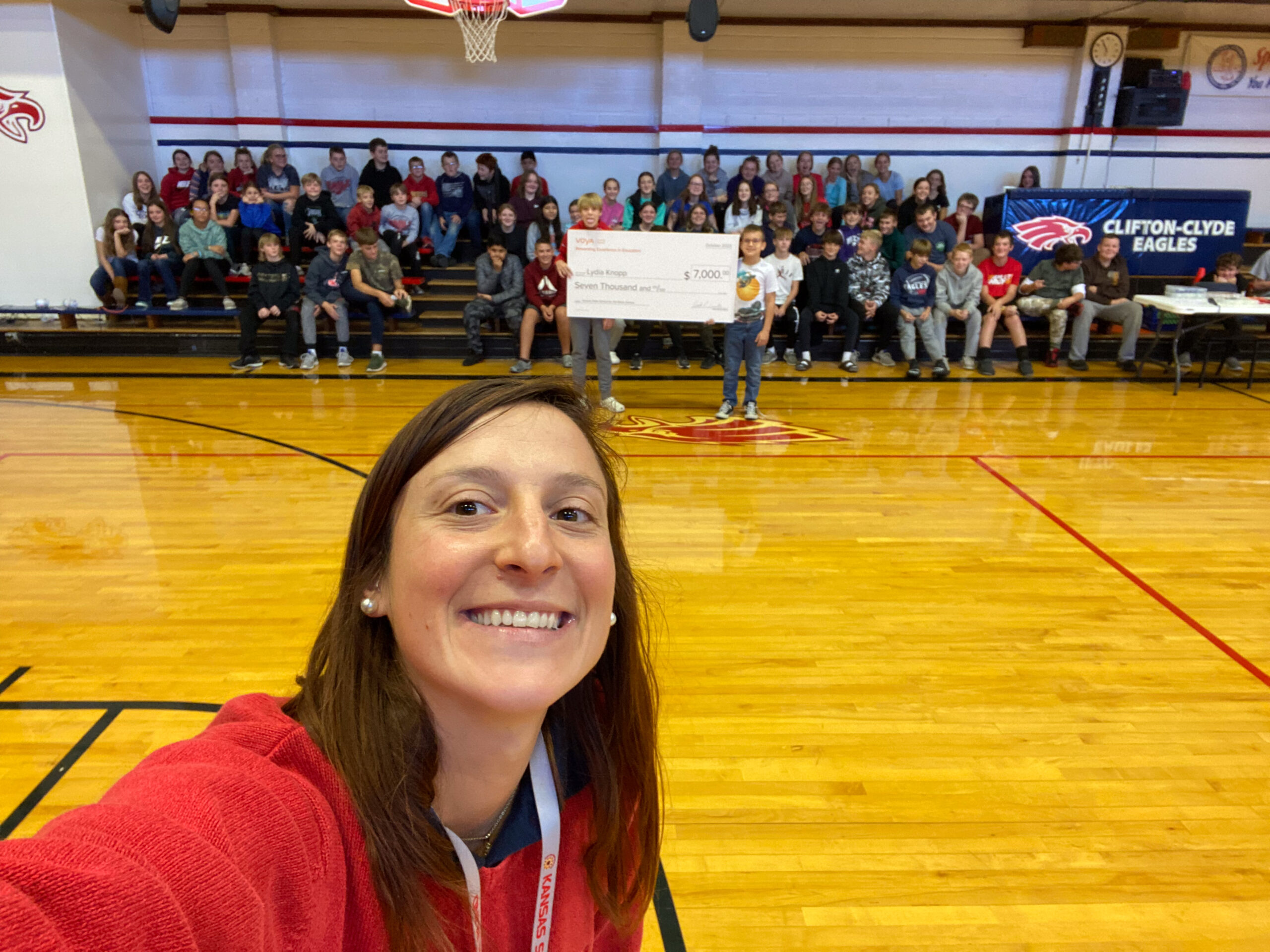 Ms. Lydia smiles while taking a selfie as a large group of students sit on gym bleachers and holds a large check