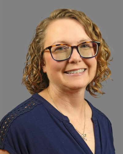 Anna Cyr has chin-length, curly blonde hair, wears glasses, a navy blue blouse and smiles for the camera.