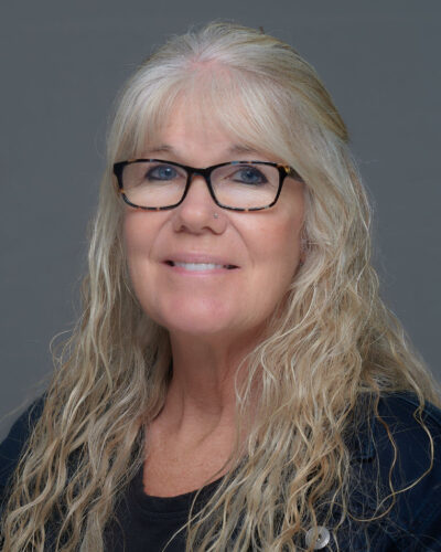 Angie Hilt has long, wavy white hair, wears glasses, a black shirt and smiles at the camera.