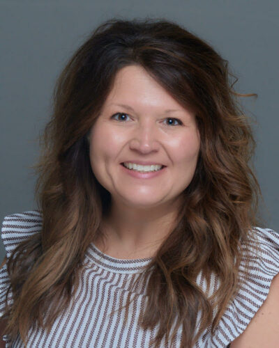 Brandi Smith has long wavy brown hair, wears a striped blue and white top and smiles at the camera.