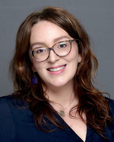 Heather Pedersen has long, reddish-brown wavy hair, wears glasses, a black blouse and smiles at the camera.