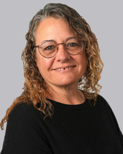 Julie Marston has curly brown hair, wears round glasses and a black sweater, and smiles for the camera.