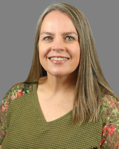 Sabrina McAdoo has long, dark grey hair, wears an olive green shirt and smiles for the camera.